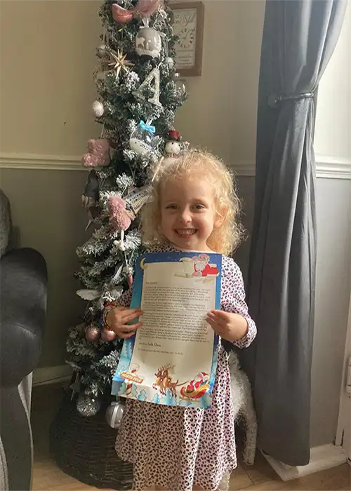 Child holding Christmas letter by decorated tree.